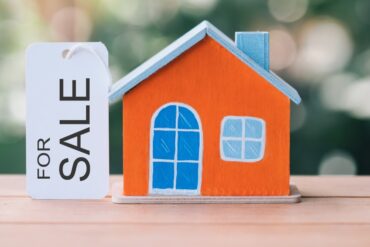 Miniature orange house with blue door and a For Sale tag next to it.