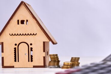 Miniature wooden house with coins on side and a calculator.