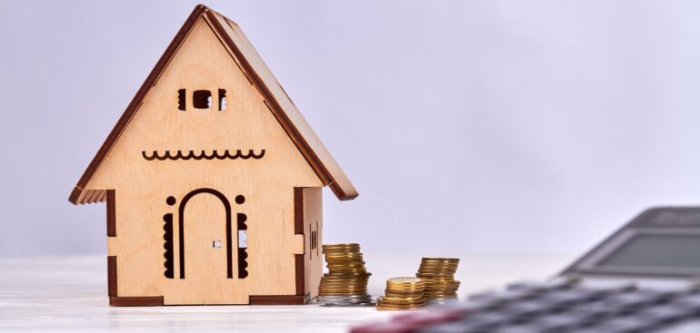 Miniature wooden house with coins on side and a calculator.